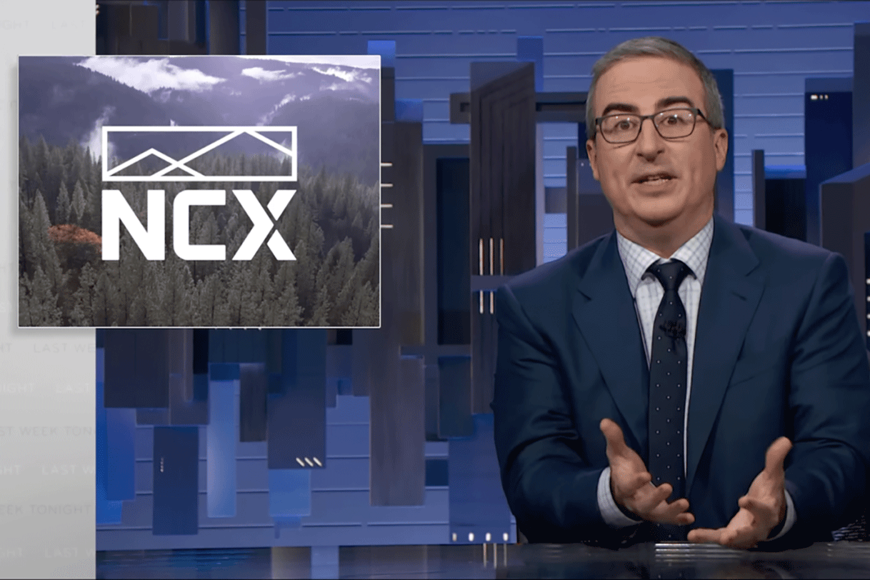 John Oliver at his desk on the show Last Week Tonight with the NCX logo appearing on the screen